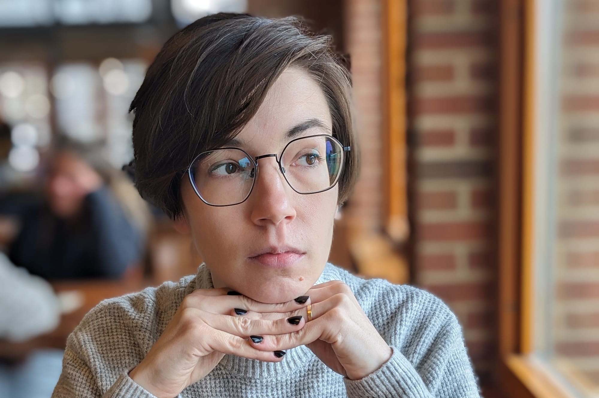 A non-binary white person with short, dark brown hair and black-rimmed glasses, resting their chin on laced fingers and looking out a window.